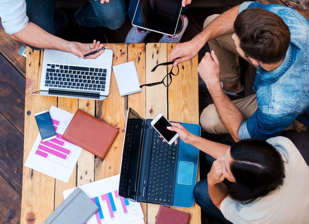 Group of young professionals sitting around a wooden table with laptops, smartphones, notebooks, and charts, collaborating on a project.