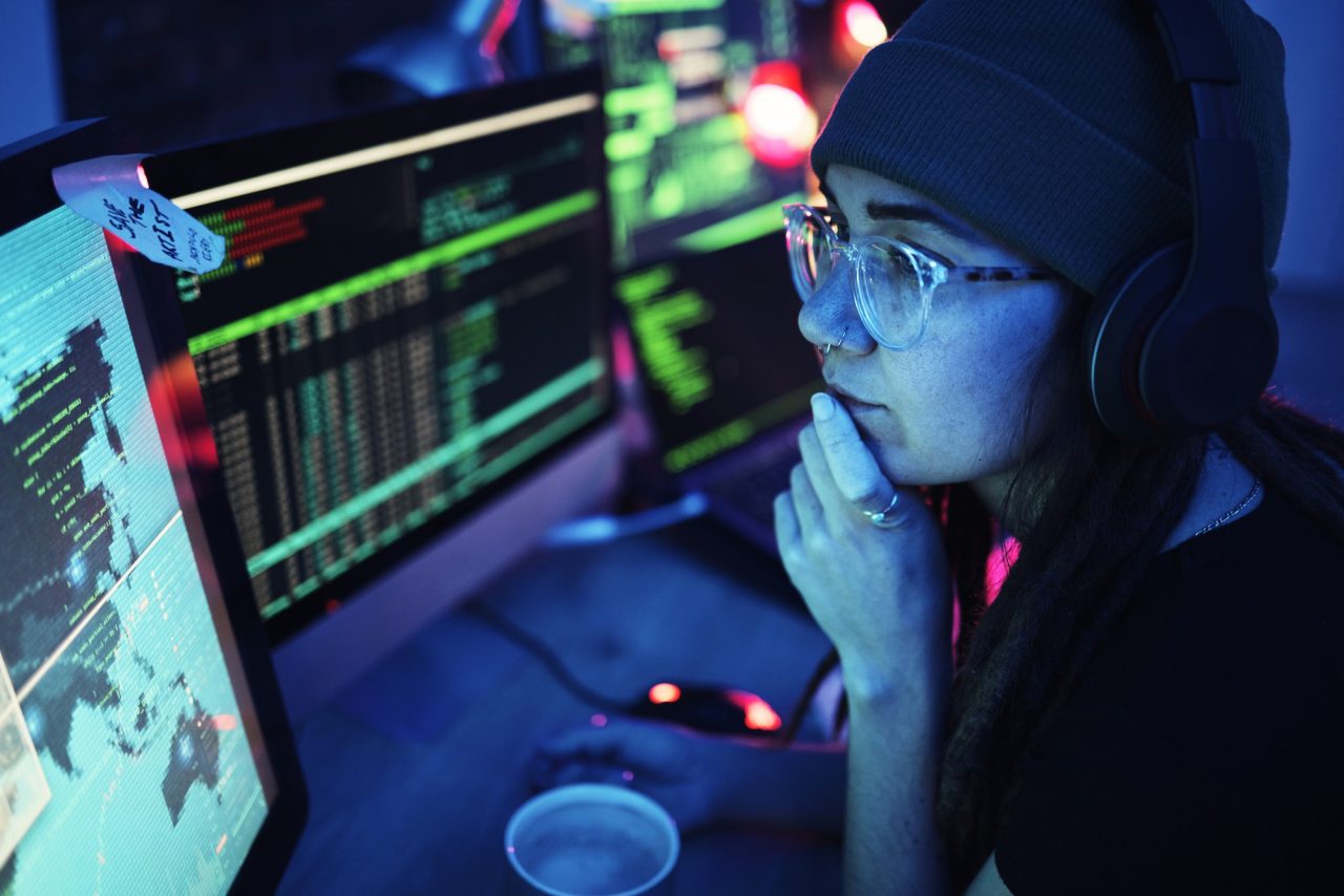 Young woman wearing glasses and headphones working on multiple computer screens with coding and data displayed in a dark room.